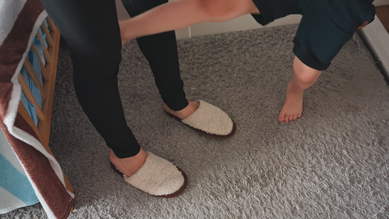 Momento della routine mattutina di mamma e figlio. Tenera scena in camera da letto con la madre che aiuta il bambino scalzo a mettere le pantofole sul tappeto accanto alla culla, con movimenti delicati, mani che guidano, illuminazione calda e accogliente, un ambiente domestico.