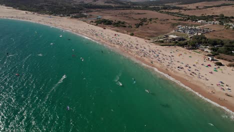 sideways-aerial-view-of-many-kitesurfers-doing-practice-and-having-fun-at-Tarifa-Beach,-Cadiz,-spain