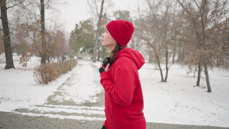 Side view of athlete wearing red beanie and jacket walking thoughtfully along snowy pathway in winter park, holding headphones with serene expression, surrounded by bare trees