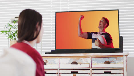 Caucasian-woman-watching-tv-with-caucasian-male-rugby-player-with-ball-on-screen