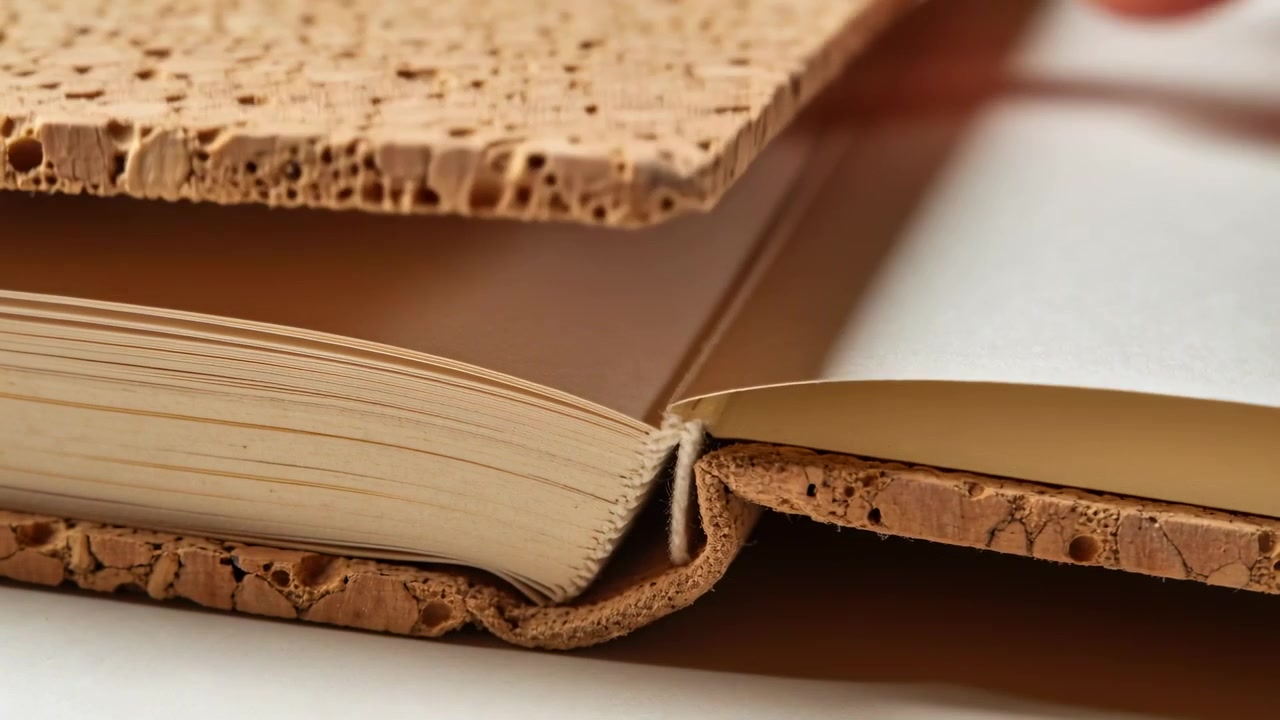 Una mano parziale che solleva la copertina rigida di sughero di un libro su un tavolo bianco, rivelando le prime pagine.