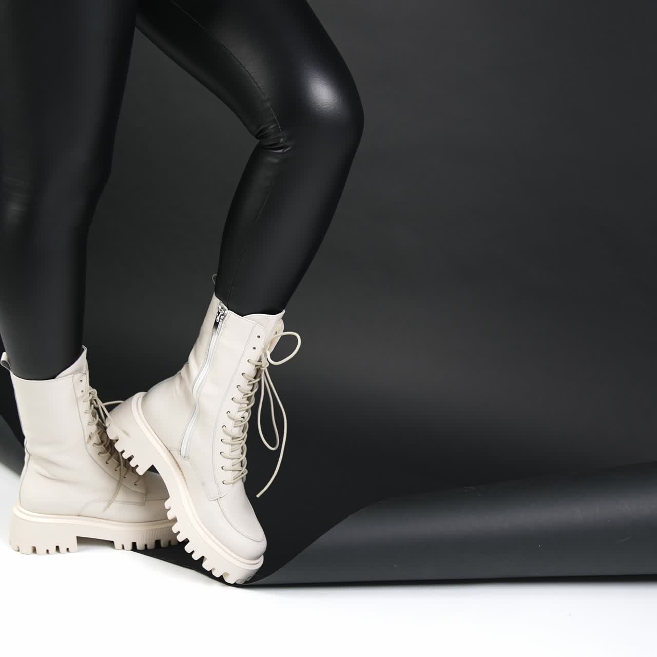 Woman in black leather leggings wears white middle-length boots with tractor soles. Model posing in studio