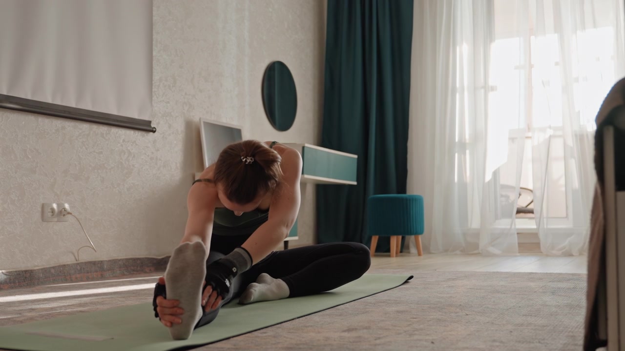 Giovane donna caucasica che esegue uno stretching dei muscoli posteriori della coscia da seduta e una flessione laterale su un tappetino in una camera da letto luminosa, respiro controllato, mobilità delicata, attenzione alla flessibilità e pratica di fitness domestico calma.