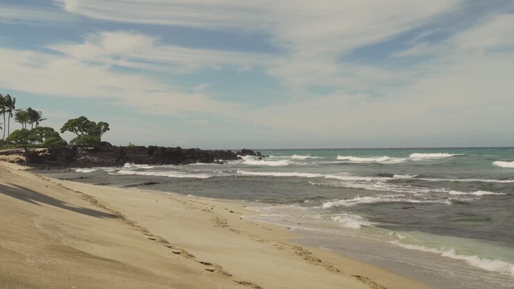 Grandes vagues balayant une plage immaculée et se brisant sur une formation rocheuse de lave au loin.
