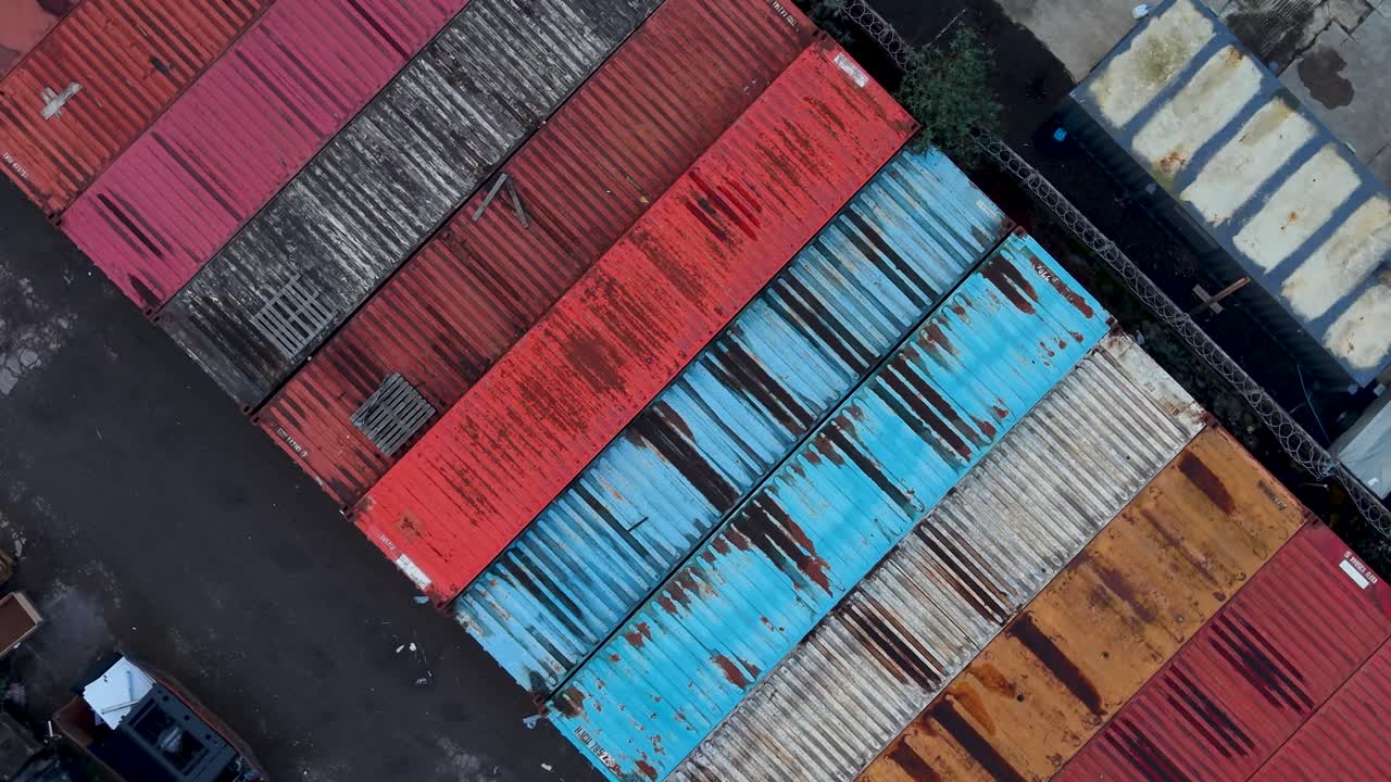 Kontainer pengiriman berwarna-warni terlihat dari drone saat berputar dan naik di atas area kargo, menampilkan kotak logam merah, oranye, hitam, putih, dan biru dalam susunan industri