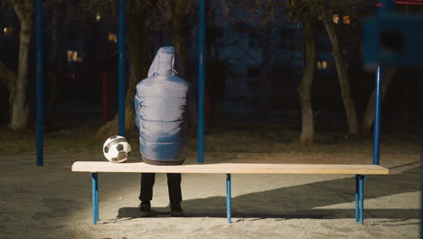 back view of a person wearing a blue hooded jacket sitting on a bench outdoor, at night with a soccer ball placed beside him illuminated by background light