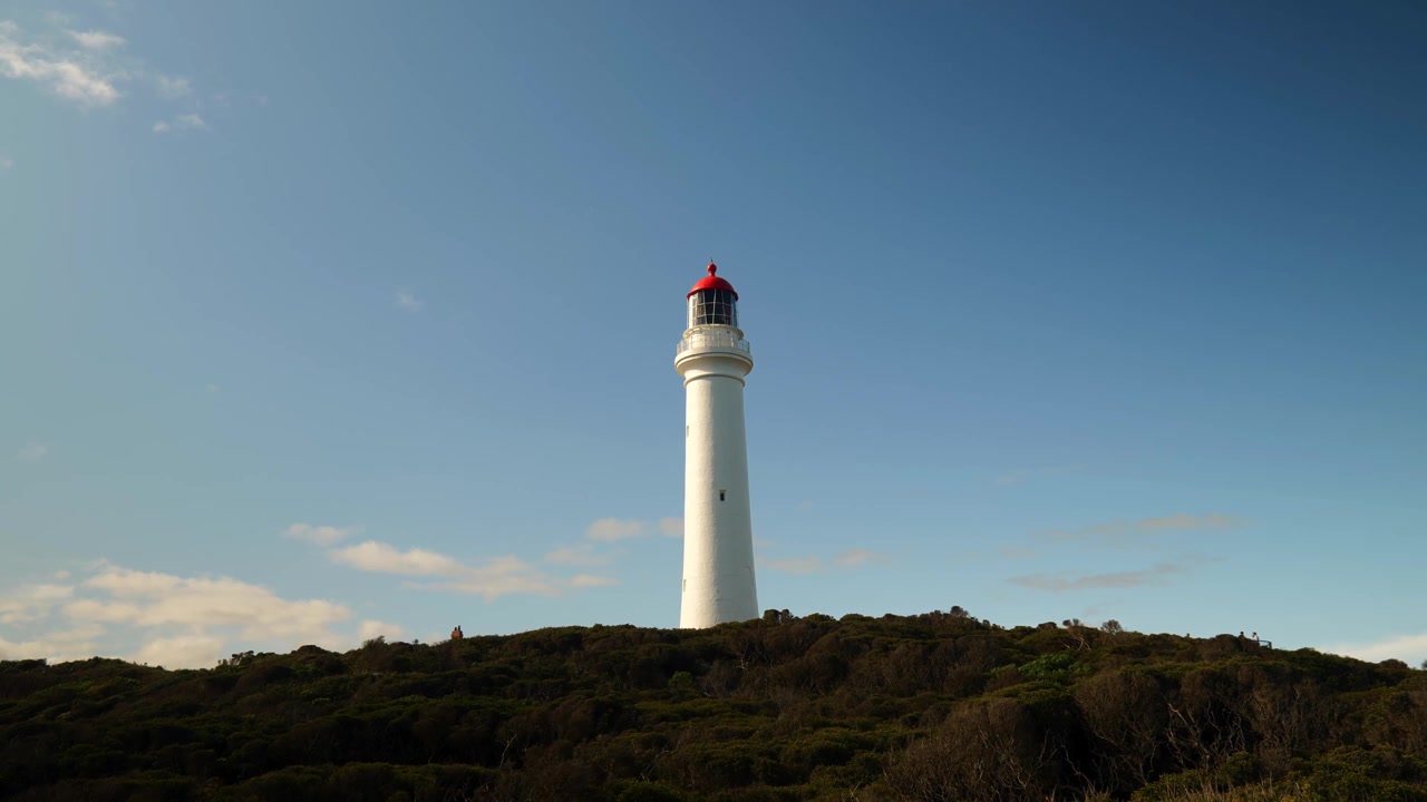 Un faro che si erge imponente su una scogliera costiera, circondato da cespugli verdi sottostanti, che si affaccia sull'oceano vasto e sulla costa frastagliata.