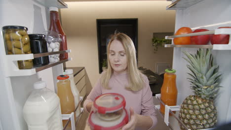 A-middle-aged-woman-takes-a-container-of-food-out-of-the-refrigerator.-View-from-inside-the-refrigerator