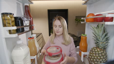 una mujer de mediana edad saca un recipiente de comida del refrigerador. vista desde el interior del refrigerador