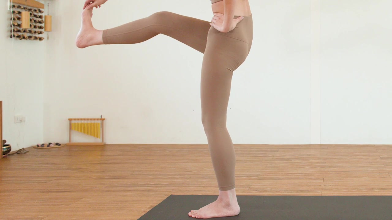 A woman performs a standing leg stretch on a yoga mat in a bright studio.