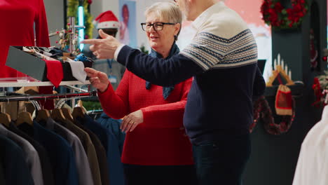 Woman-browsing-through-xmas-adorn-clothing-store-racks-with-husband-in-Christmas-shopping-spree.-Elderly-couple-searching-for-elegant-attire-garments-as-gift-for-son-during-winter-holiday-season