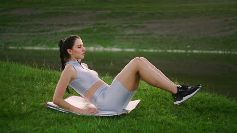 a woman does exercises for the abs muscles lying on the grass in a park near the lake lifting her body and legs. work with abs muscles. slim beautiful figure. fitness marathon