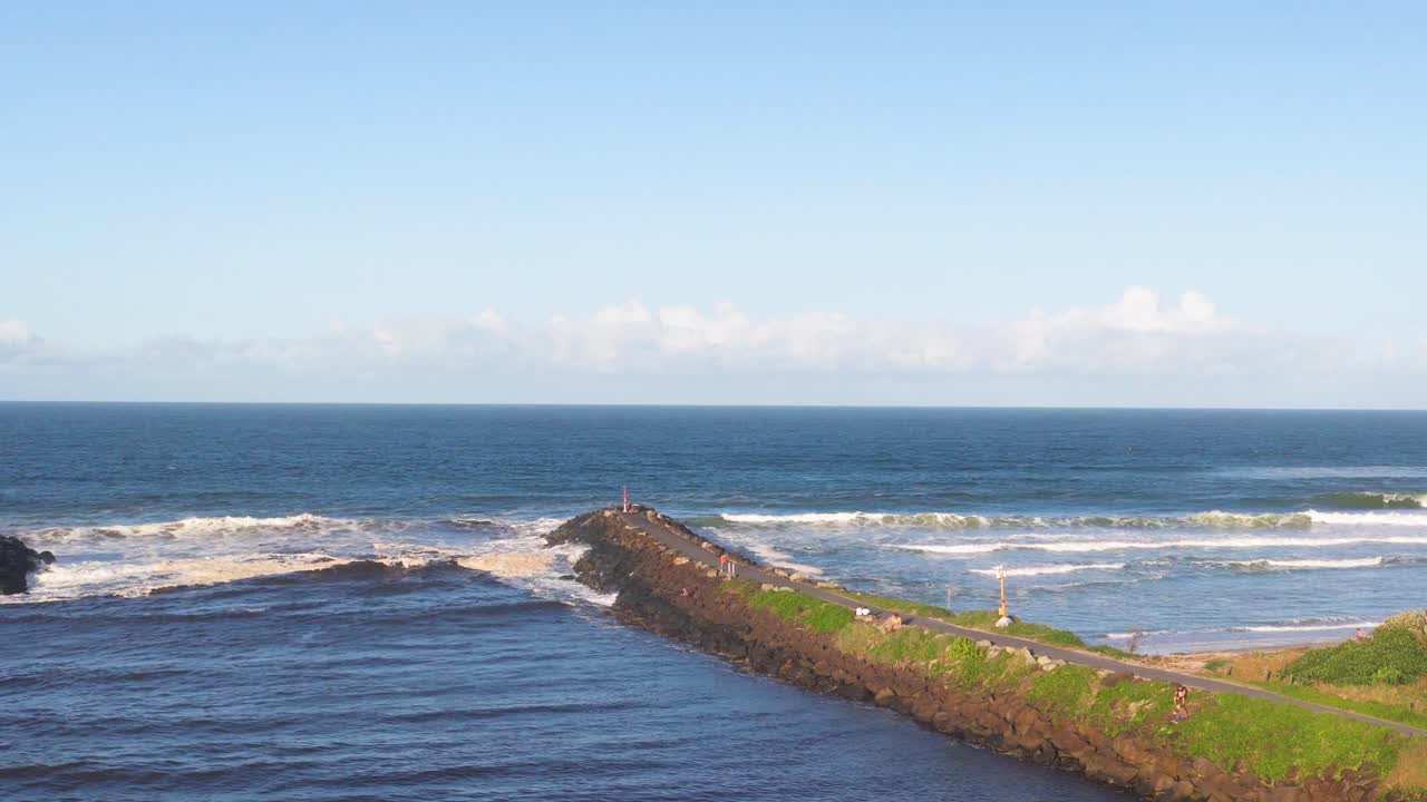 Aerial footage captures ocean waves crashing against a rocky jetty under clear skies at Brunswick Heads, Australia