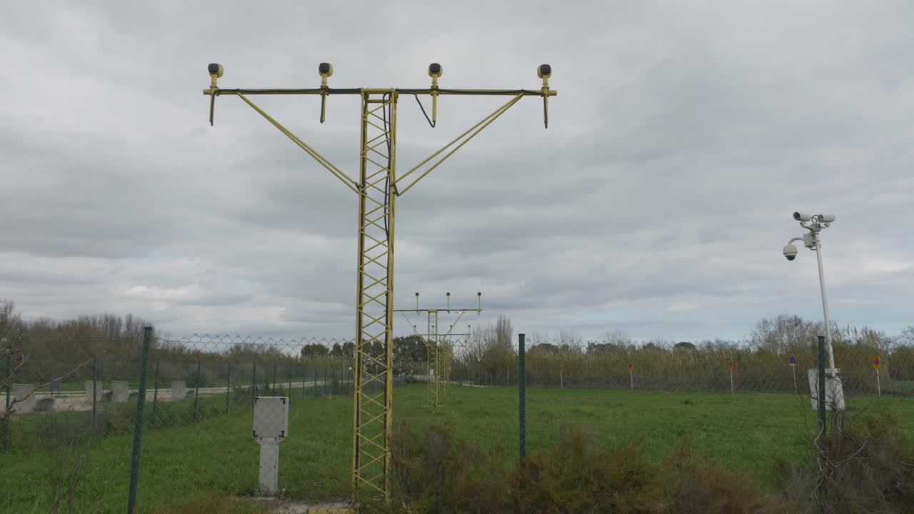 Passenger aircraft approaching runway, descending over illuminated airport infrastructure and green landscape with security monitoring cameras against cloudy sky backdrop