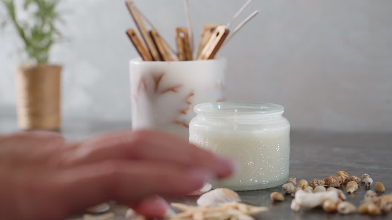 Hand adjusting arrangement of seashells and snail shells on marble surface with white decorative candle jar and incense sticks in background
