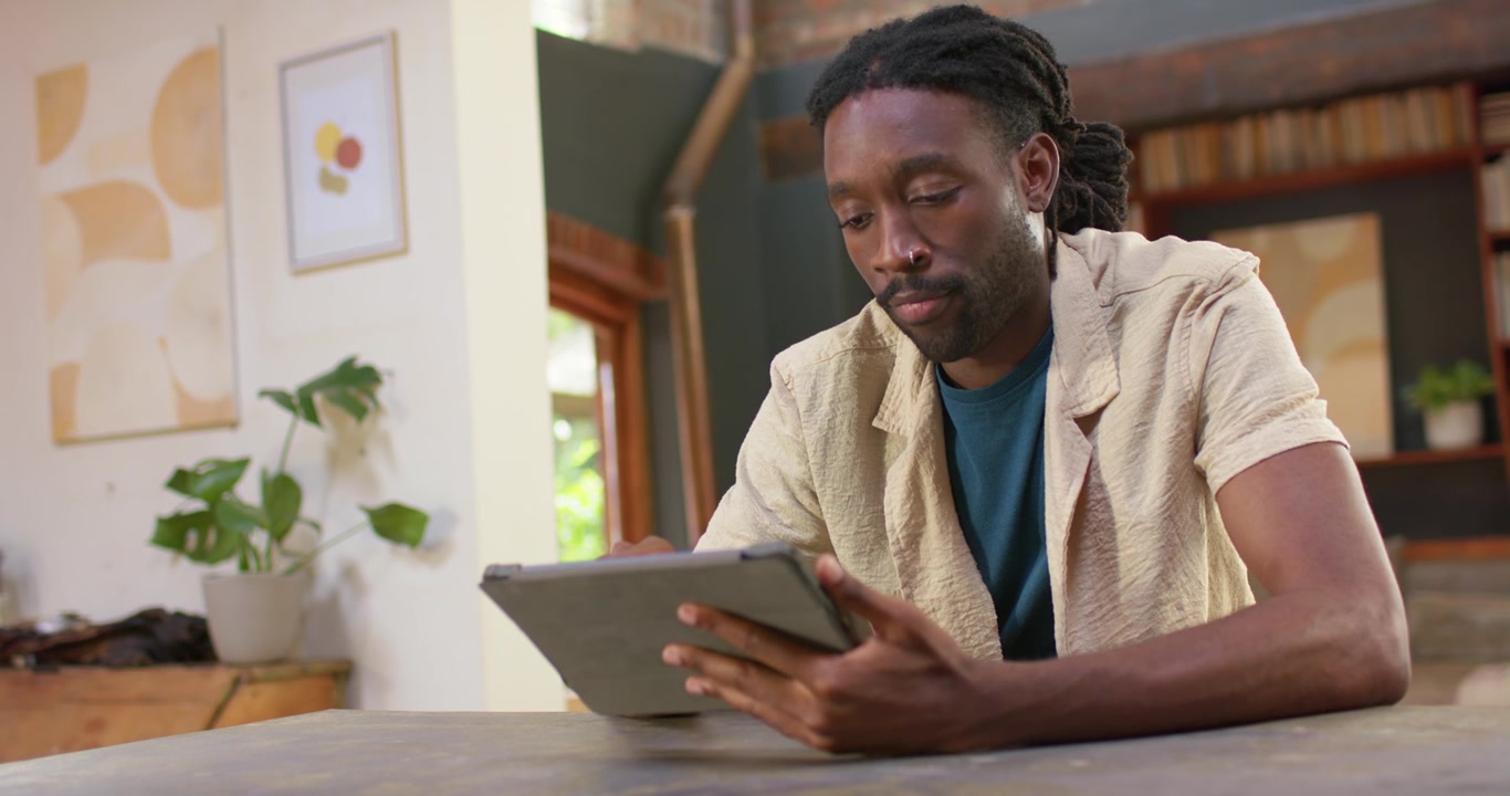 African adult male seeing tablet sitting at wooden table home holding tablet tapping reviewing work