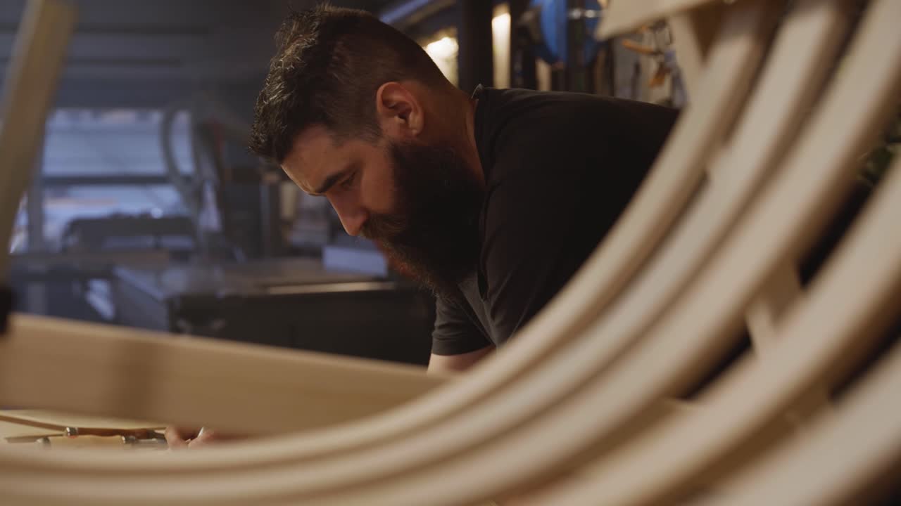 A craftsman concentrates on his woodworking project in a modern studio, seen through curved wooden pieces, symbolizing focus, craftsmanship, and artistic precision