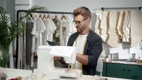 man tailor in glasses sitting at the table with a sewing machine and using tablet