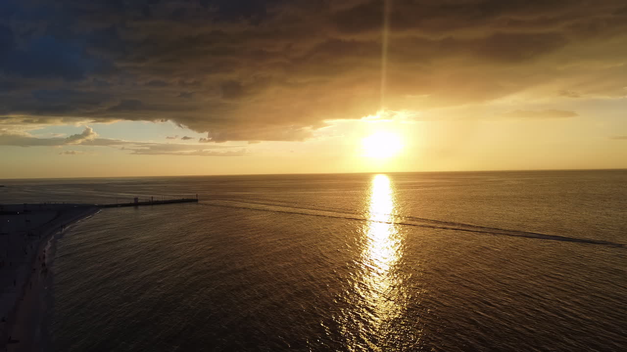 Aerial drone view of a dramatic golden sunset over the ocean along the Mexican coastline. A small boat crosses the calm sea leaving a long wake beneath colorful clouds and warm evening light