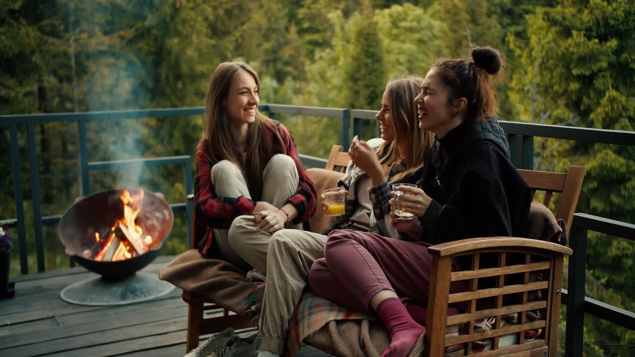 Three girls are chatting and laughing while sitting on a sofa near the barbecue in a country house overlooking the forest and mountains
