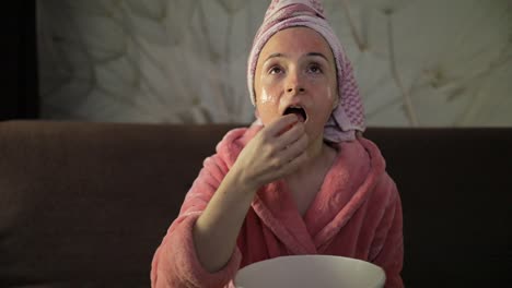 woman watching a late night movie at tv, eating popcorn. bathrobe, facial mask