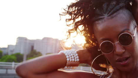 close up of young black woman wearing sunglasses plying with her hair, looking to camera on a london street at the golden hour