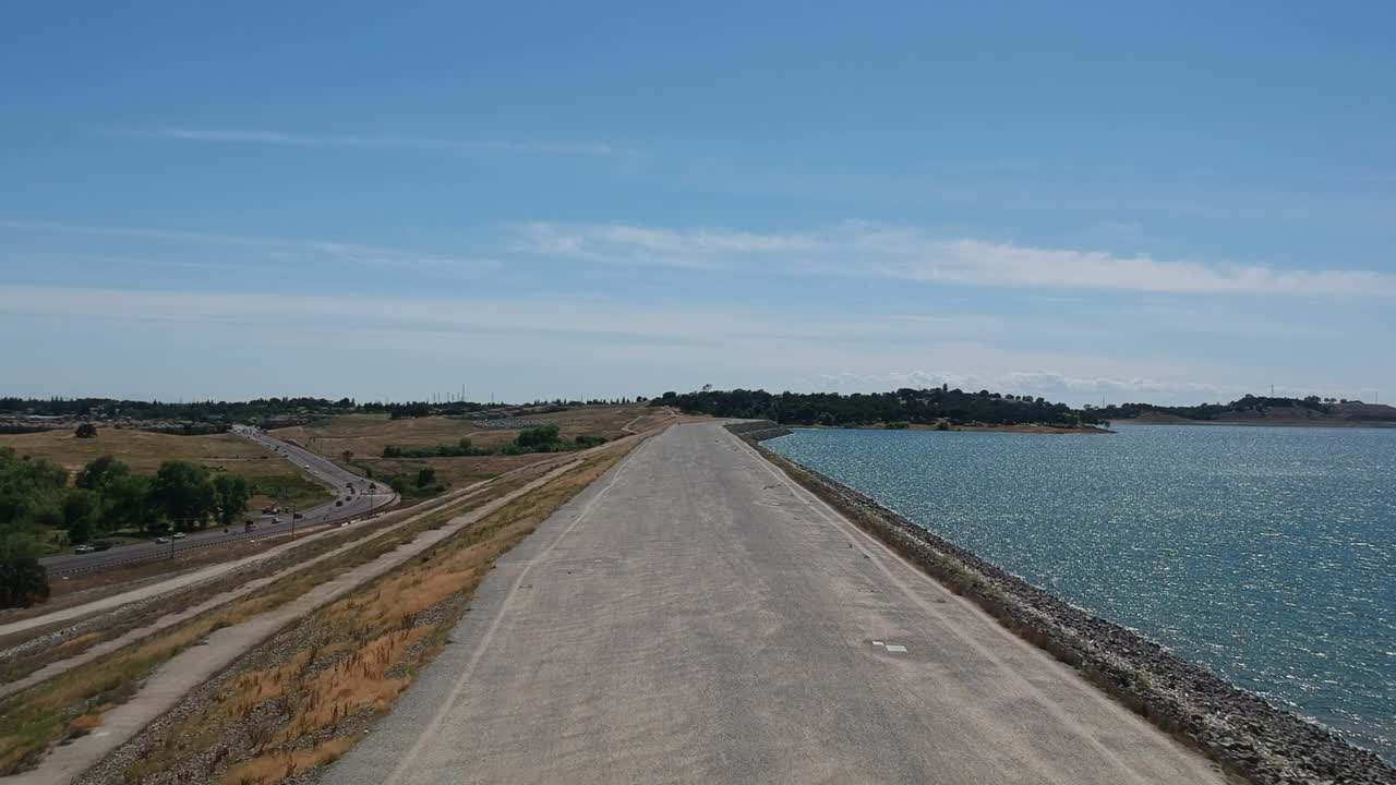오른쪽으로 폴섬 호수의 맑고 푸른 물과 초원, 왼쪽으로 도로가 있는 부드러운 포장도로에서 이륙하는 드론 영상, 푸른 하늘로 수직으로 올라가는 경치