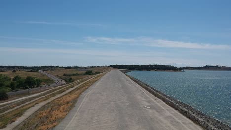 drone footage taking off from smooth pavement with crystal clear blue water of folsom lake to the right and grassland and a road to the left, with scenic views as it ascends vertically into blue sky