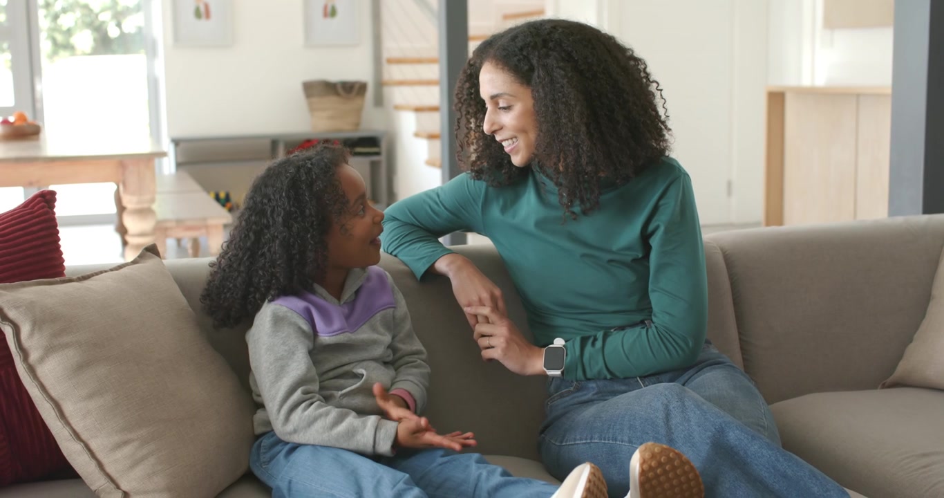 African American female mother and daughter talking on sofa at home, mother leaning, nudging child