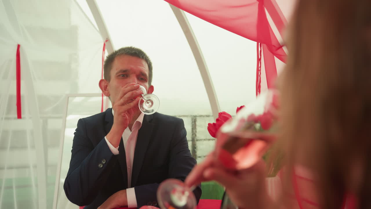 Hombre caucásico brindando con vino, cita romántica. Un hombre de negocios con traje azul marino levanta una copa para brindar con su pareja, un ramo de tulipanes rojos en un jarrón sobre la mesa, vino rosado espumoso, una sonrisa amable, luz solar tenue