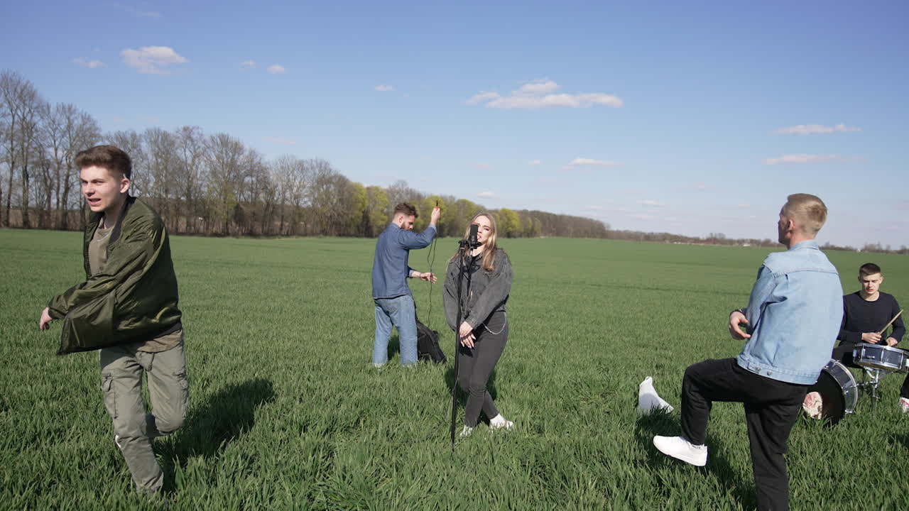Energetic and cheerful musicians perform music on green field. Young band singing and dancing outdoors. Men and a woman play drums and sing a song into a microphone.