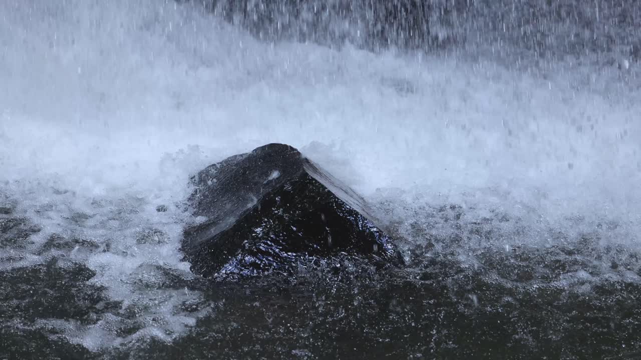 A dark rock stands in a flowing stream as a waterfall crashes over it, creating dynamic splashes. Natural lighting, steady camera, close-up view