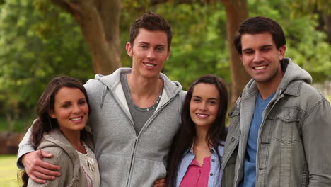 man holds his three friends and looks at the camera as they stand in a park