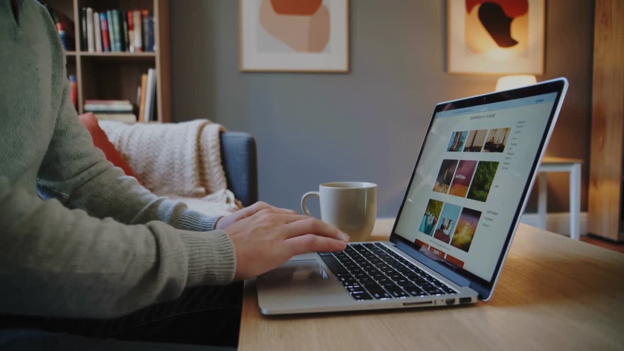 Opening laptop placing mug, person typing scrolling thumbnails reviewing images at home table