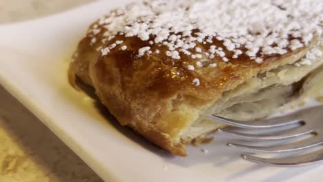 close-up of a golden brown, flaky puff pastry dessert, generously dusted with powdered sugar, a fork delicately lifts a bite, revealing the soft, airy interior, ready to be savored