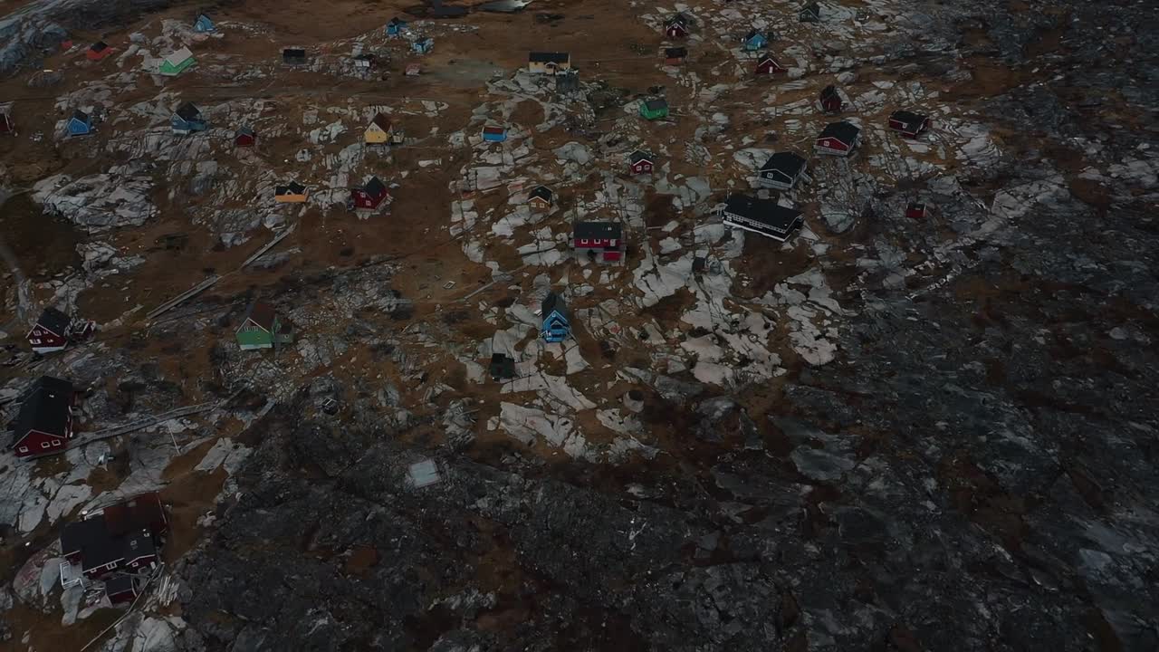 Aerial drone view of the remote Arctic village of Ittoqqortoormiit in Greenland, showing colorful houses along rocky coast with large icebergs floating in the calm Arctic sea