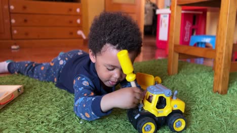 cute and expresive 3-year-old black child playing at home to fix his yellow toy bulldozer laying on a green rug