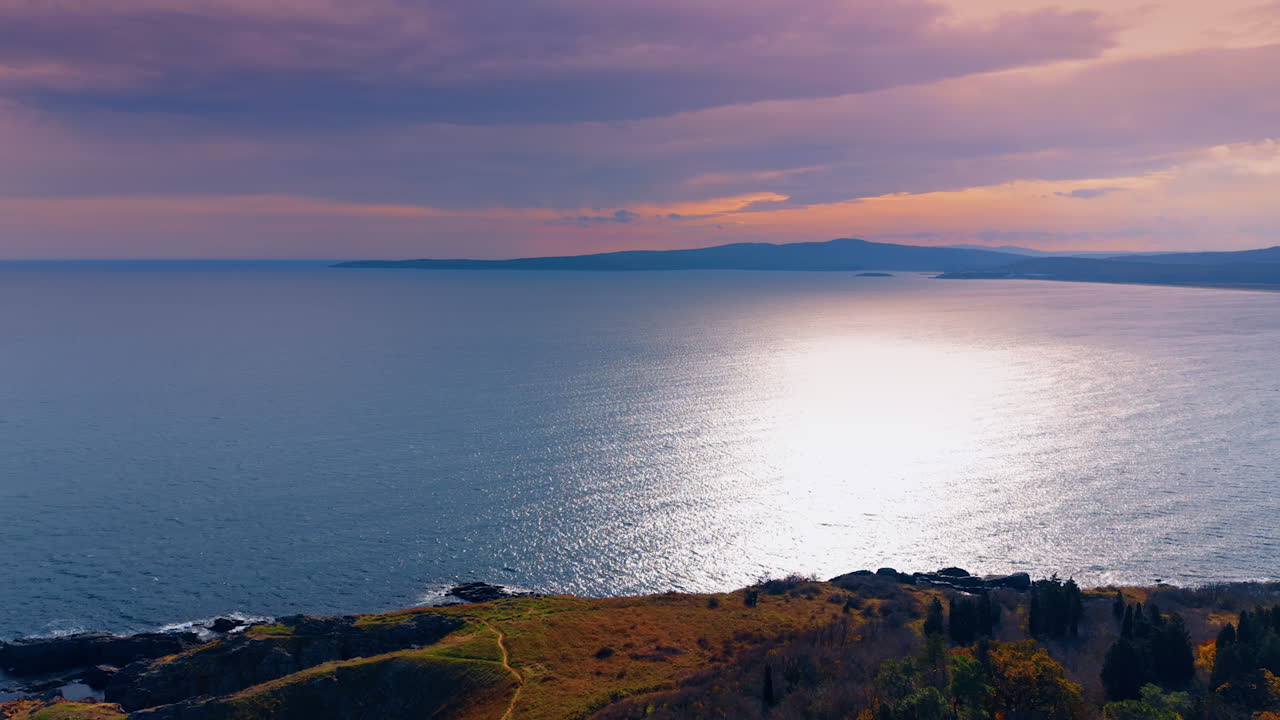A view of the coastline during sunset shows water reflecting light with dark clouds above. The distant mountains add depth to the horizon. The scene captures the transition from day to night