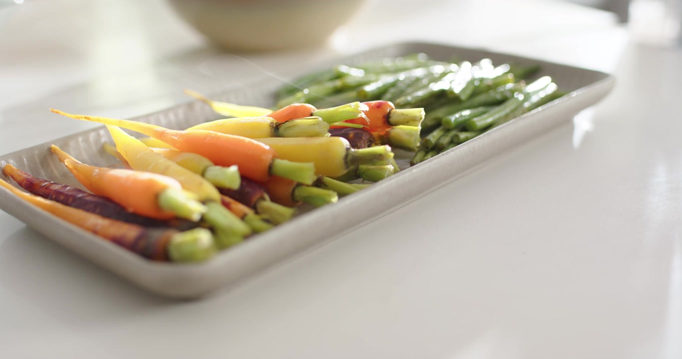 Camera pulling in, racking focus on tray of carrots and stalks on white counter, showing freshness