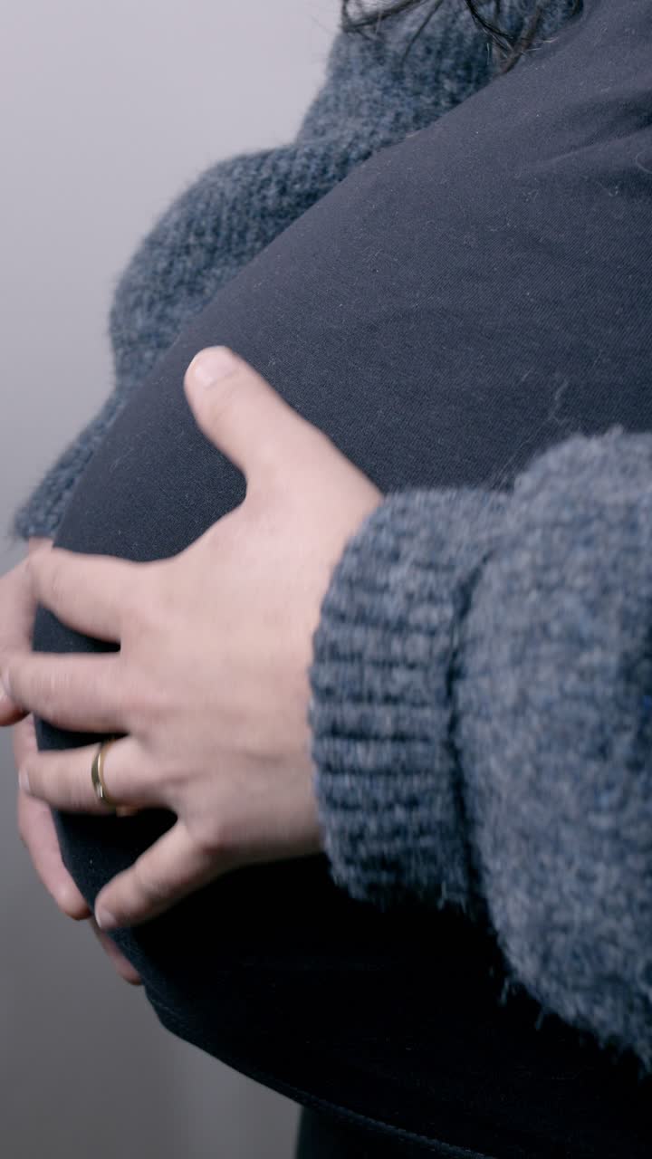 A tender close-up shot of a pregnant woman's hands gently resting on her belly. The cozy sweater adds warmth to this intimate vertical video, perfect for mobile viewing