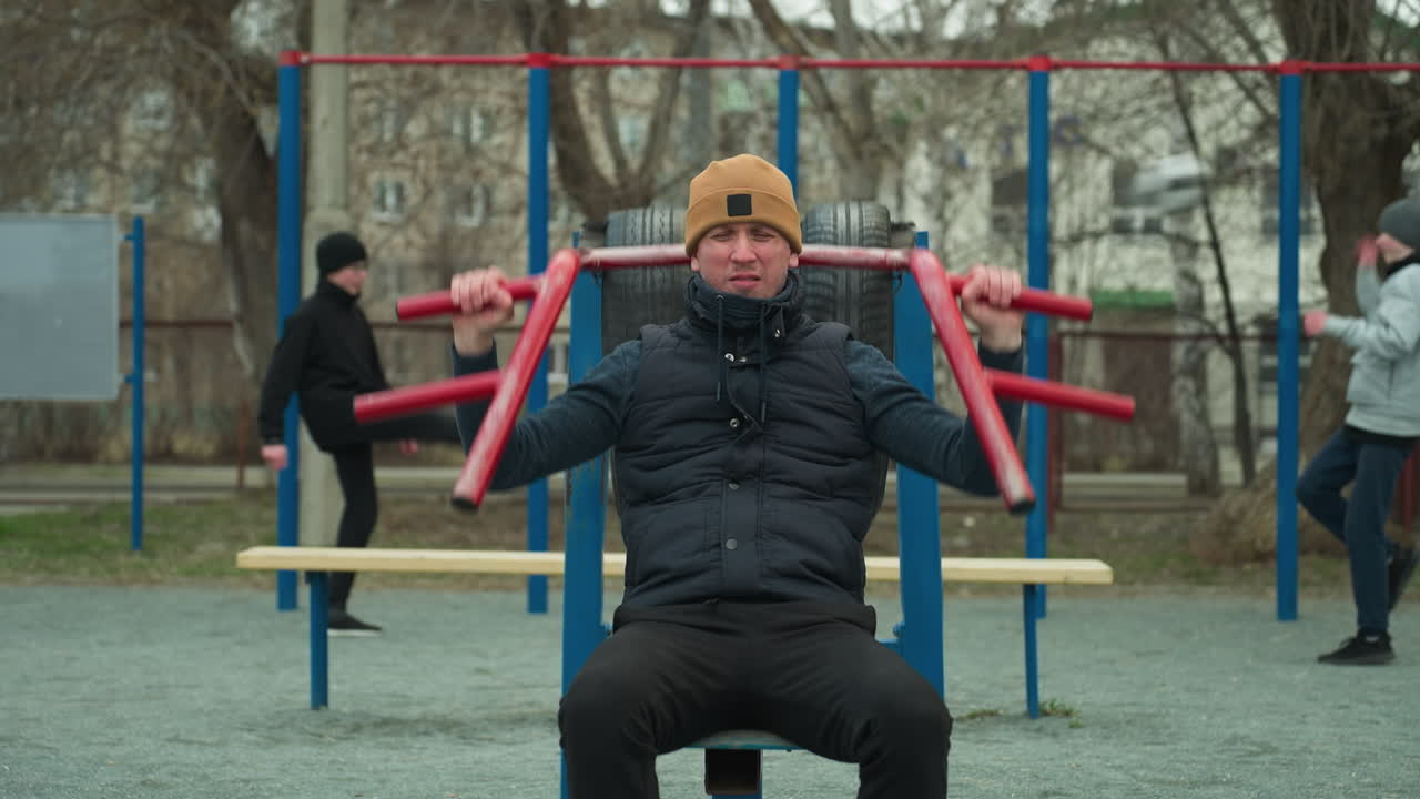 A coach wearing a brown beanie and black jacket works out on outdoor equipment, lifting red bars above his head and down, in the background, two boys are playing football