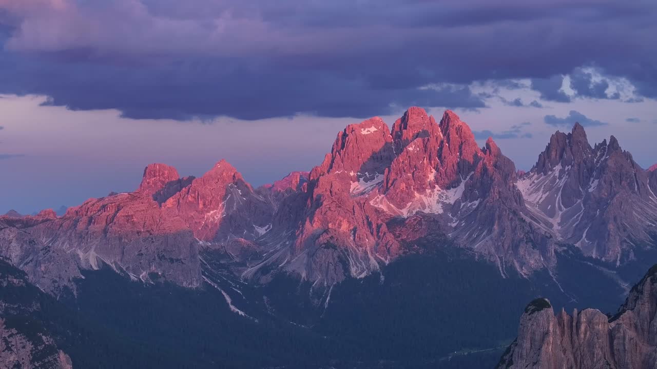 Bagliore alpino al tramonto sulle Tre Cime (Drei Zinnen) con sfumature rosa e arancioni che illuminano le vette montane, Dolomiti Italia, ripresa aerea con teleobiettivo.