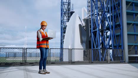 vista lateral completa del cuerpo de una ingeniera asiática con casco de seguridad trabajando en una computadora portátil mientras está de pie con el transbordador espacial