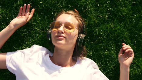 Chica-Escuchando-Música-En-Streaming-Con-Auriculares-En-Verano-En-Un-Prado.-Césped-Verde.-Chica-Sonriente-Con-Gafas-De-Sol-Amarillas-Y