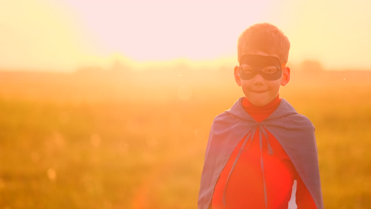 un niño vestido como un superhéroe de pie con una máscara y una capa roja viendo la puesta de sol en el campo en el verano. noche de verano el niño sueña y hazañas heroicas y cómics