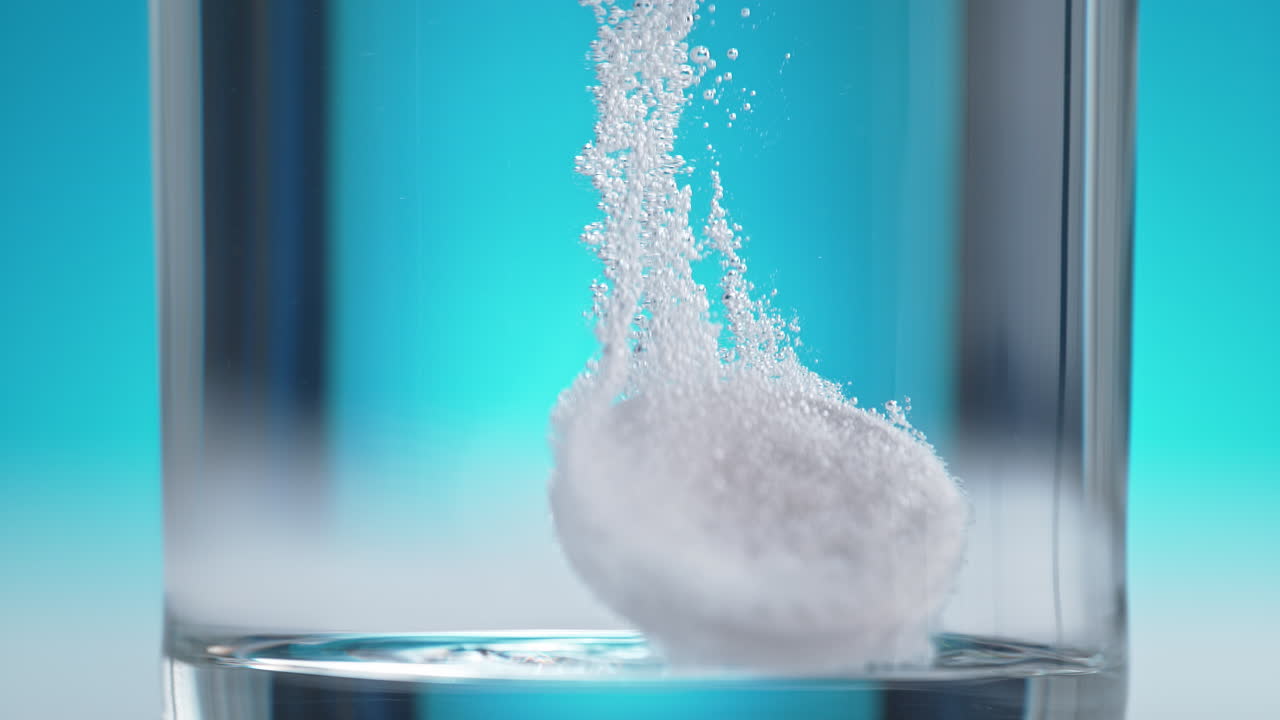 la tableta de aspirina se hunde en el fondo del vaso, humeando y burbujeando en macro y cámara lenta en el fondo azul