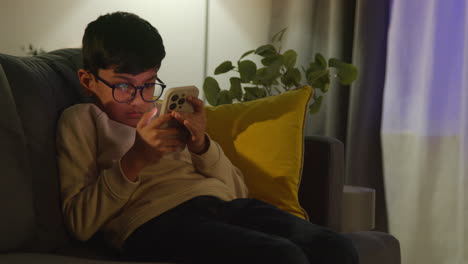 young boy sitting on sofa in lounge at home playing game on mobile phone at night