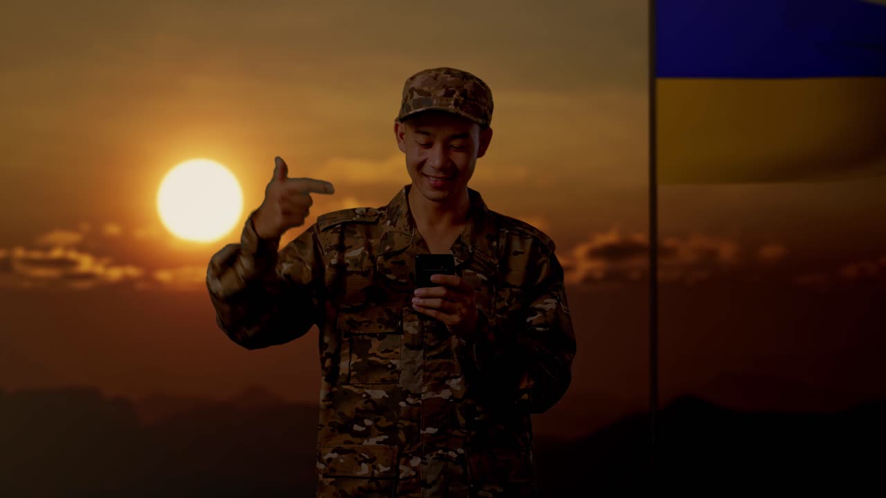 soldado sonriente en uniforme de camuflaje con teléfono inteligente al atardecer cerca de la bandera ucraniana