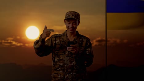 soldado sonriente en uniforme de camuflaje con teléfono inteligente al atardecer cerca de la bandera ucraniana
