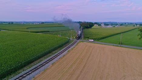 Una-Vista-Aérea-De-Un-Tren-De-Vapor-Que-Se-Acerca-Volando-Viajando-A-Través-De-Tierras-De-Cultivo-Y-Campos-De-Maíz-Que-Soplan-Humo-En-Un-Día-Soleado-De-Verano